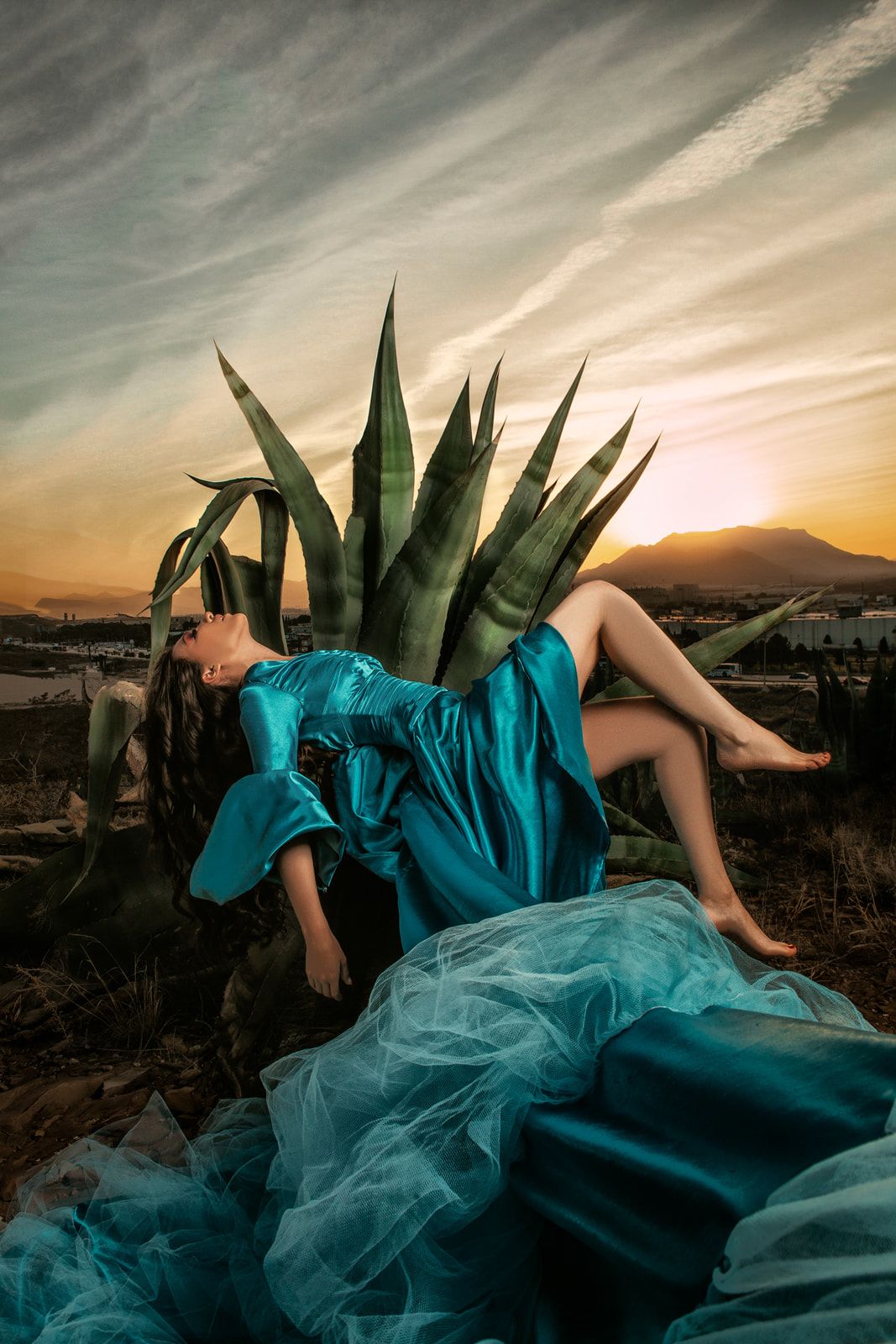 Artistic portrait with turquoise dress and agave plant against sunset landscape.