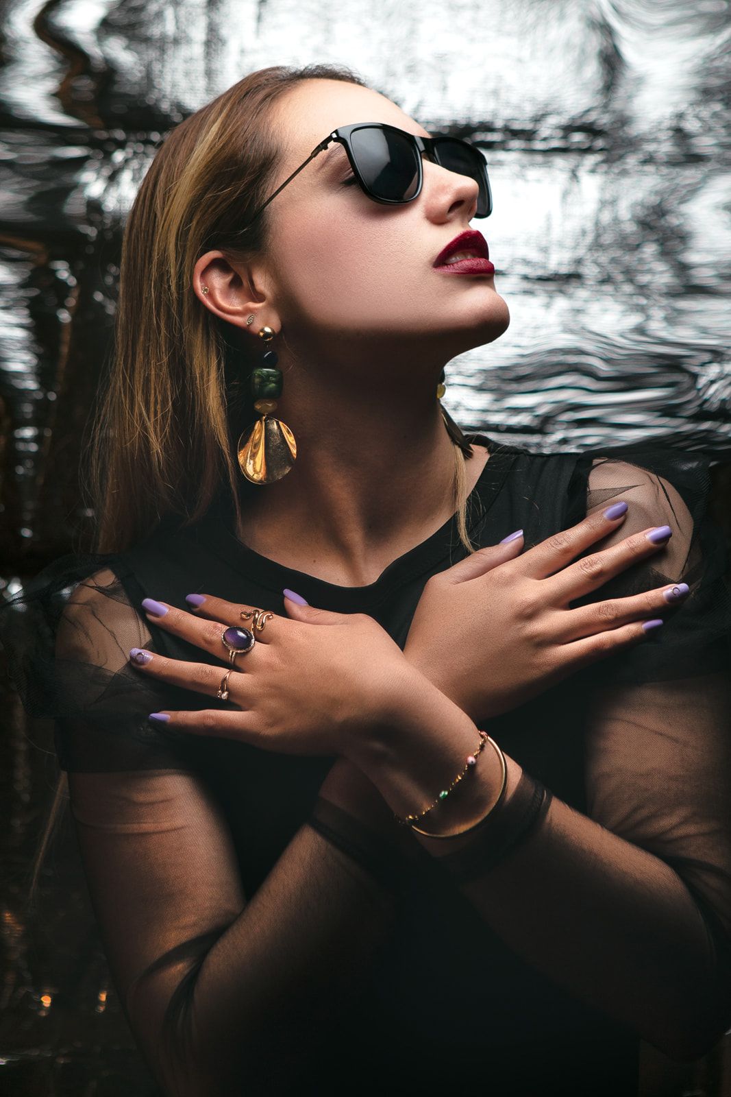 Dramatic fashion portrait with sunglasses and jewelry against textured dark background.