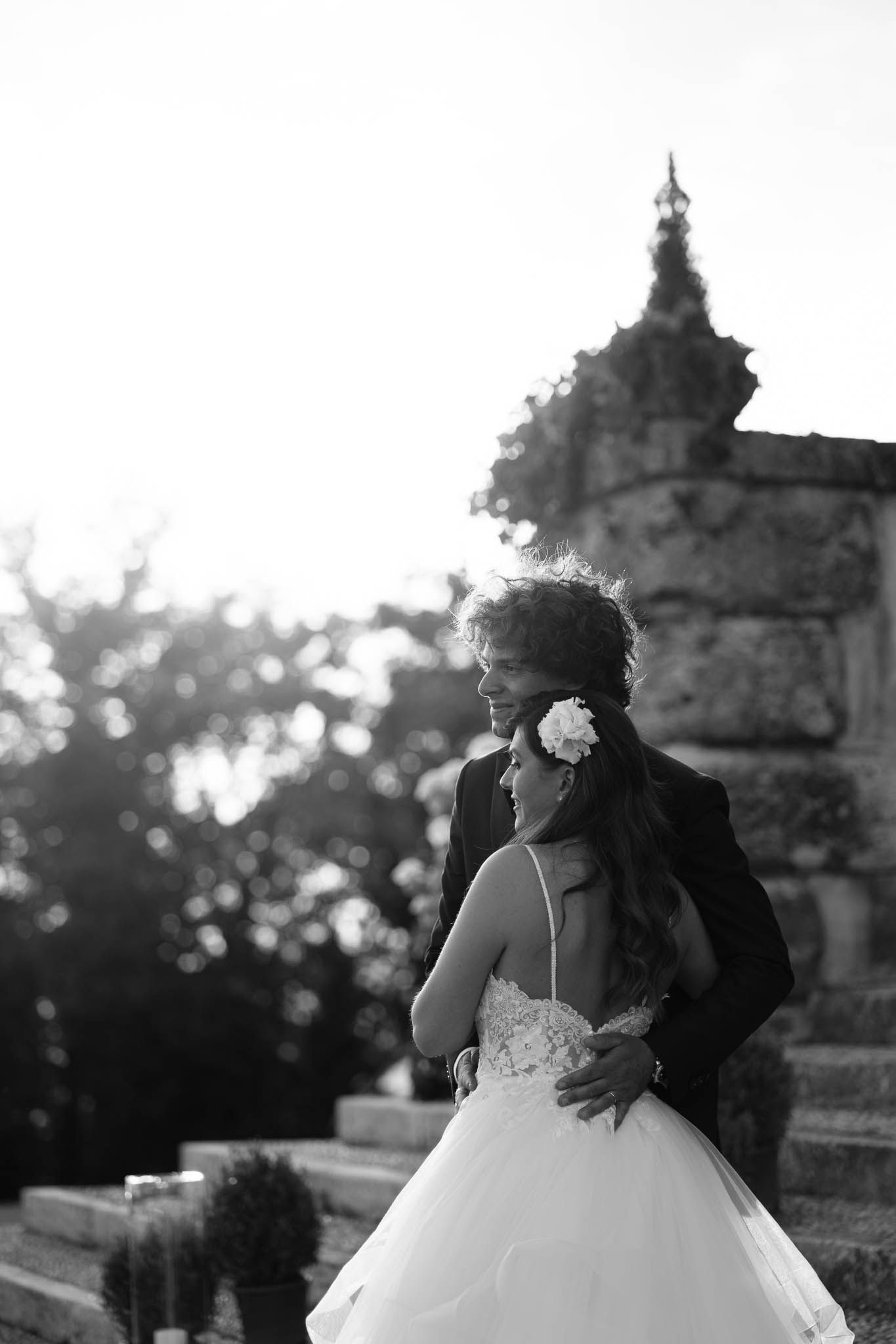 A romantic black and white wedding portrait against a stone building at sunset.
