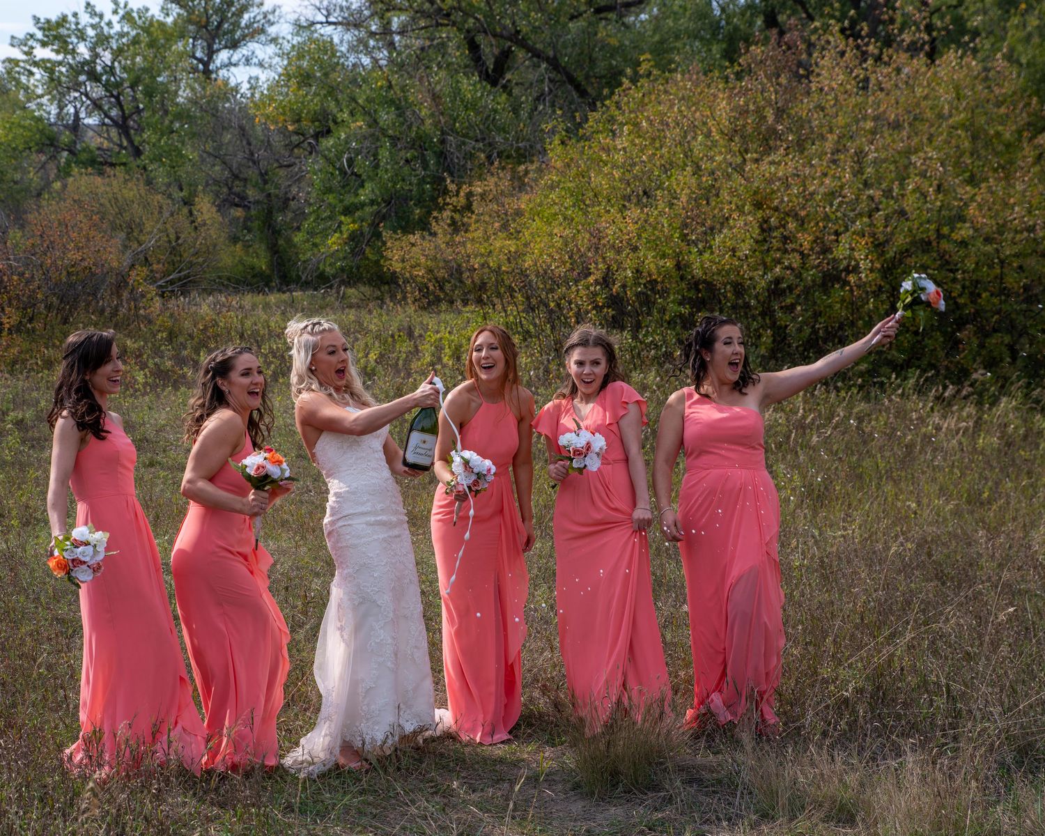 Bridesmaids in coral dresses pose with bride in outdoor nature setting.