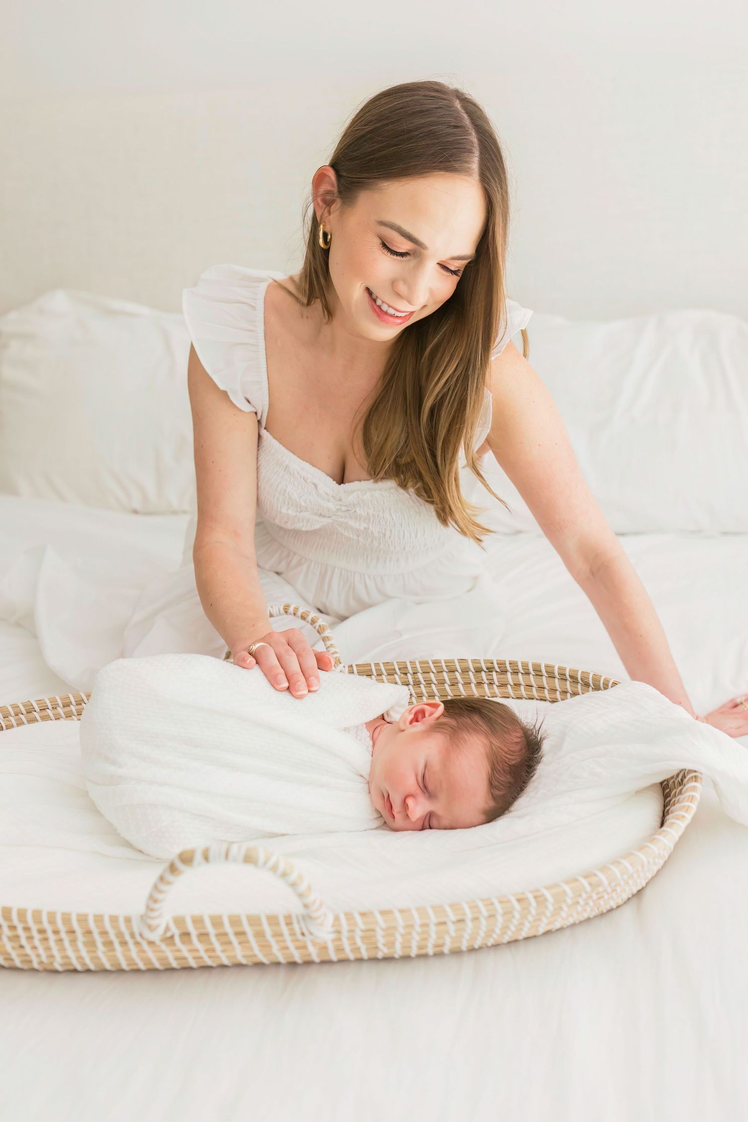 A mother in a white dress lovingly watches over her sleeping newborn baby in a woven bassinet on white bedding.