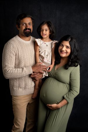 a family of three with one on the way stand in a studio smiling for family photos