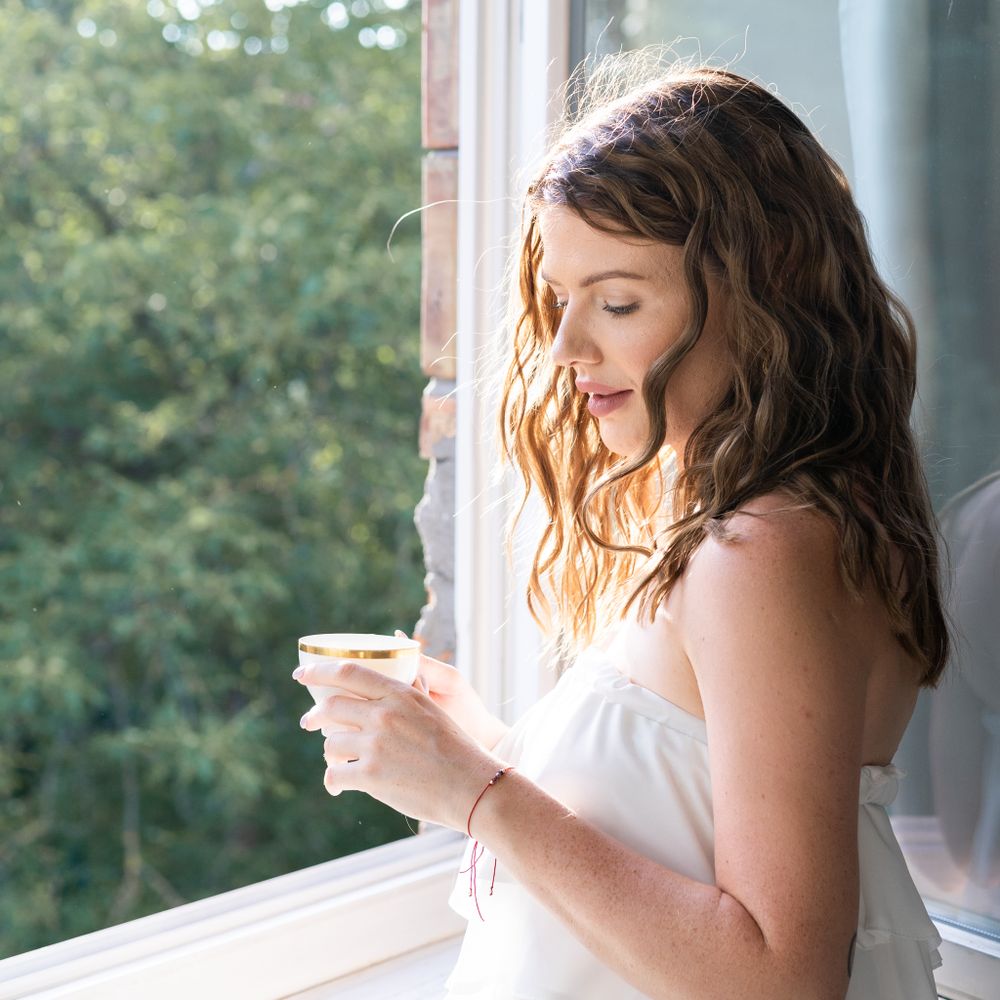 Person in white dress looks peacefully out window while holding coffee cup.