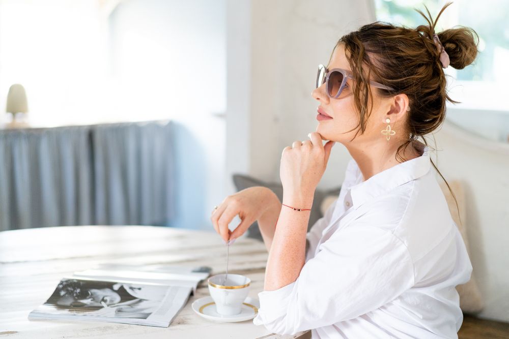 Someone in white shirt and sunglasses sits thoughtfully at table with magazine.