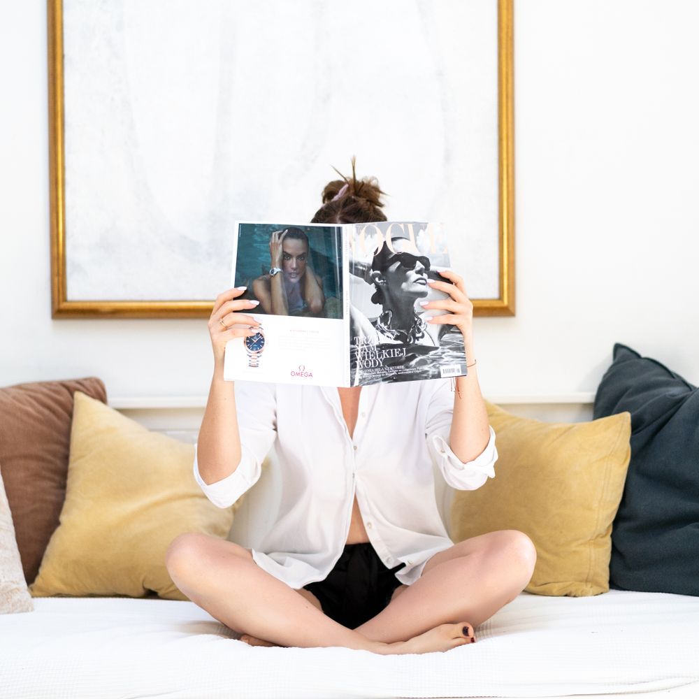 Person in white shirt sits cross-legged on couch holding magazines.