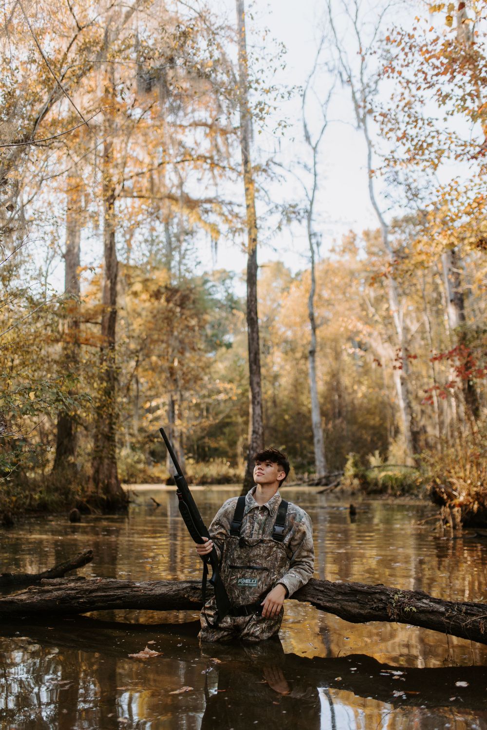 Hunter in camouflage gear sitting on fallen log in swampy autumn forest.