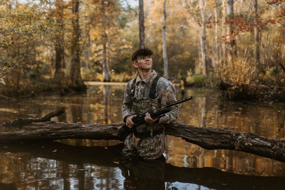 Hunter in camouflage gear standing in autumn swamp water with hunting equipment.