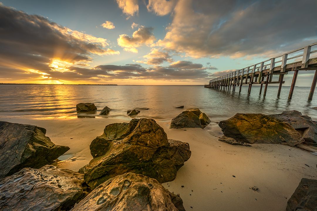 Wooden pier extends into ocean at sunset with mossy rocks in foreground.
