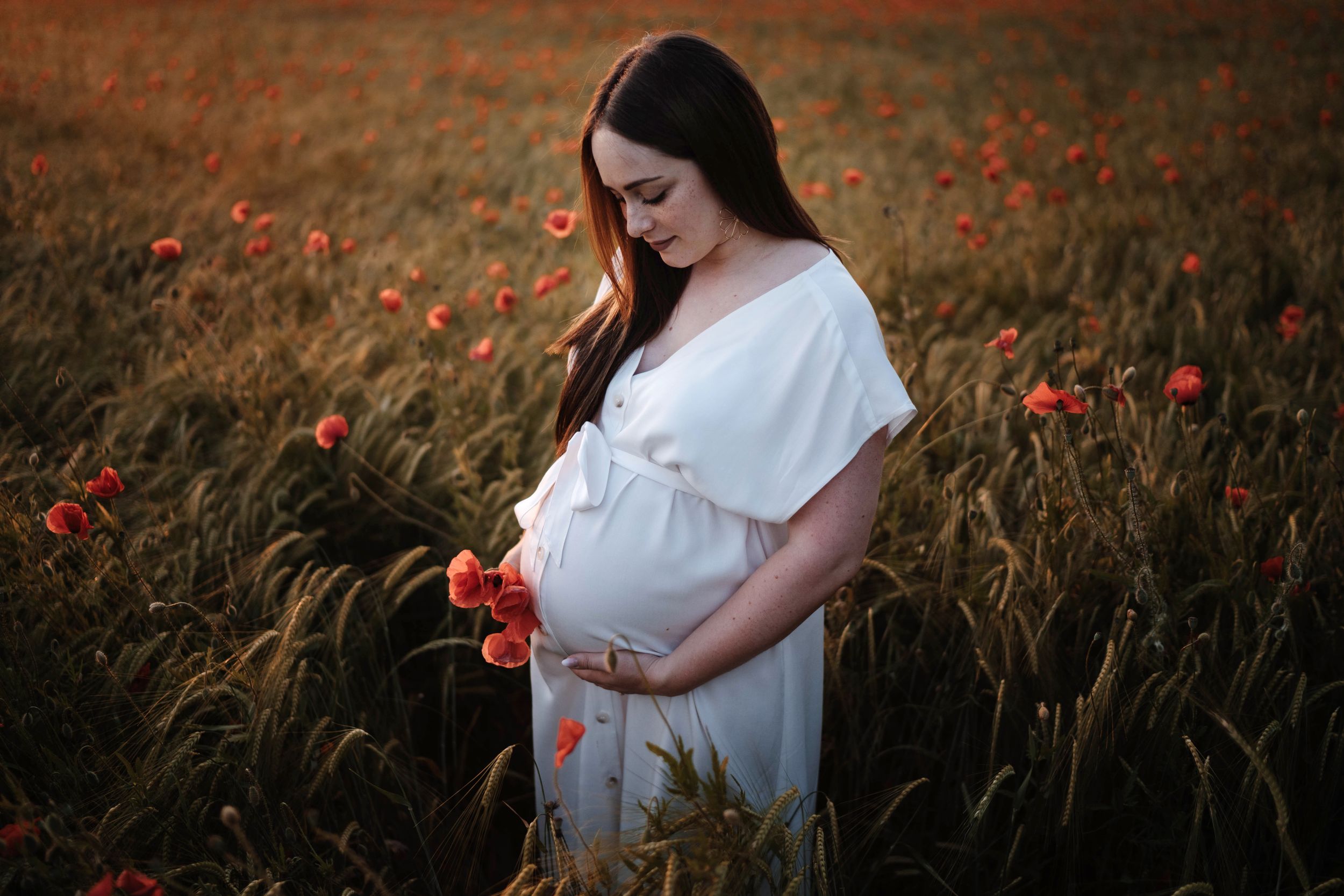 A person in a white dress stands in a golden wheat field dotted with vibrant red poppies at sunset.
