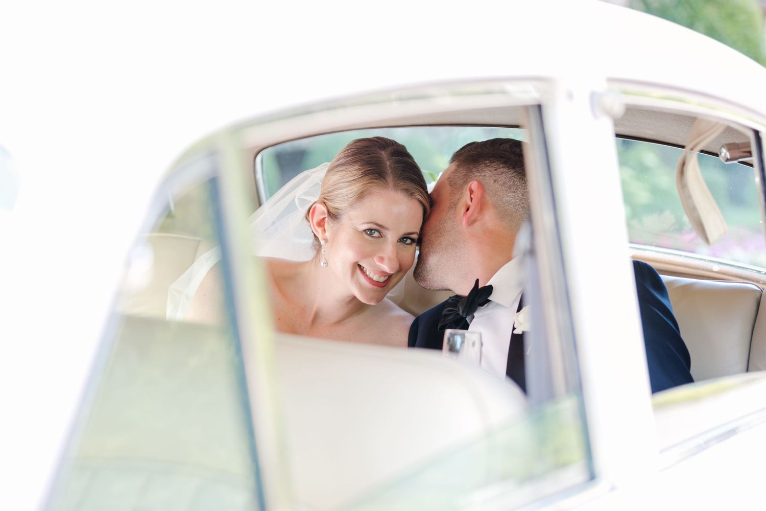 A romantic wedding photo captured through a vintage car window showing a tender moment between newlyweds.