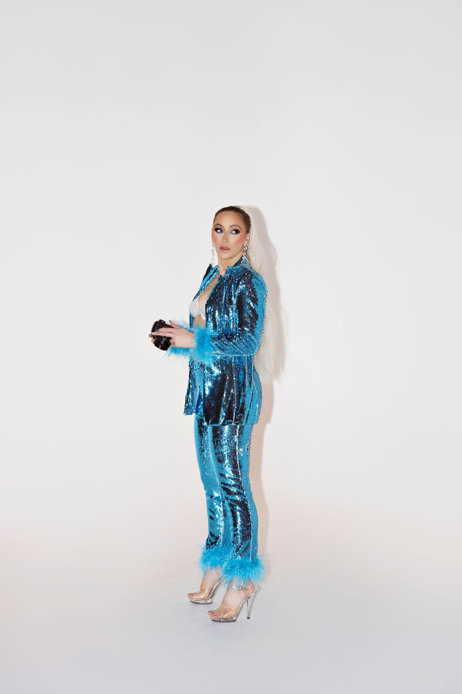 Model wearing a glittery blue sequined pantsuit and silver heels poses against white backdrop.
