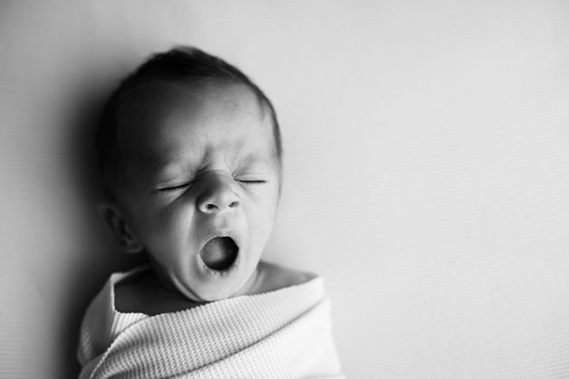 Yawning newborn baby wrapped in white blanket in black and white portrait.