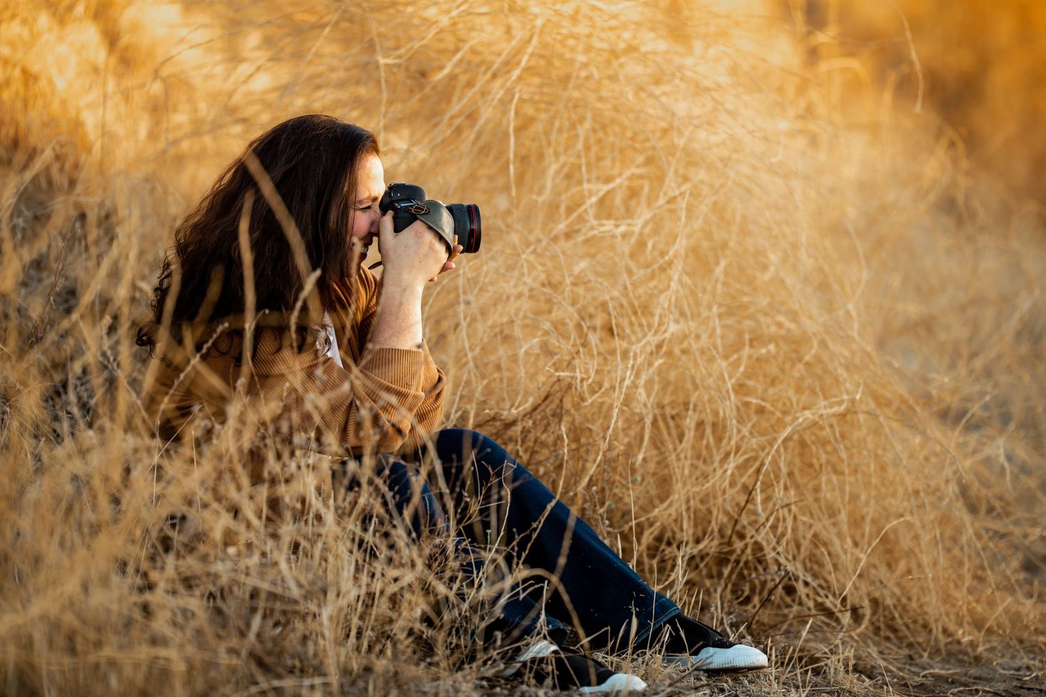 Photographer with long dark hair sitting among golden grass taking photos with a DSLR camera.