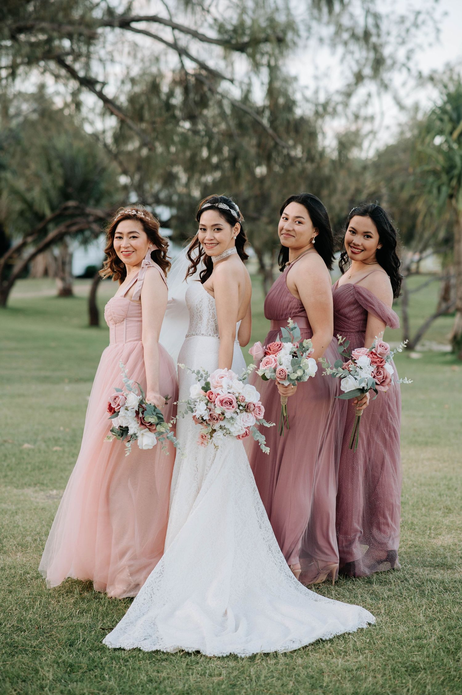 Bride in white gown with bridesmaids in pink dresses hold bouquets while posing on grass.