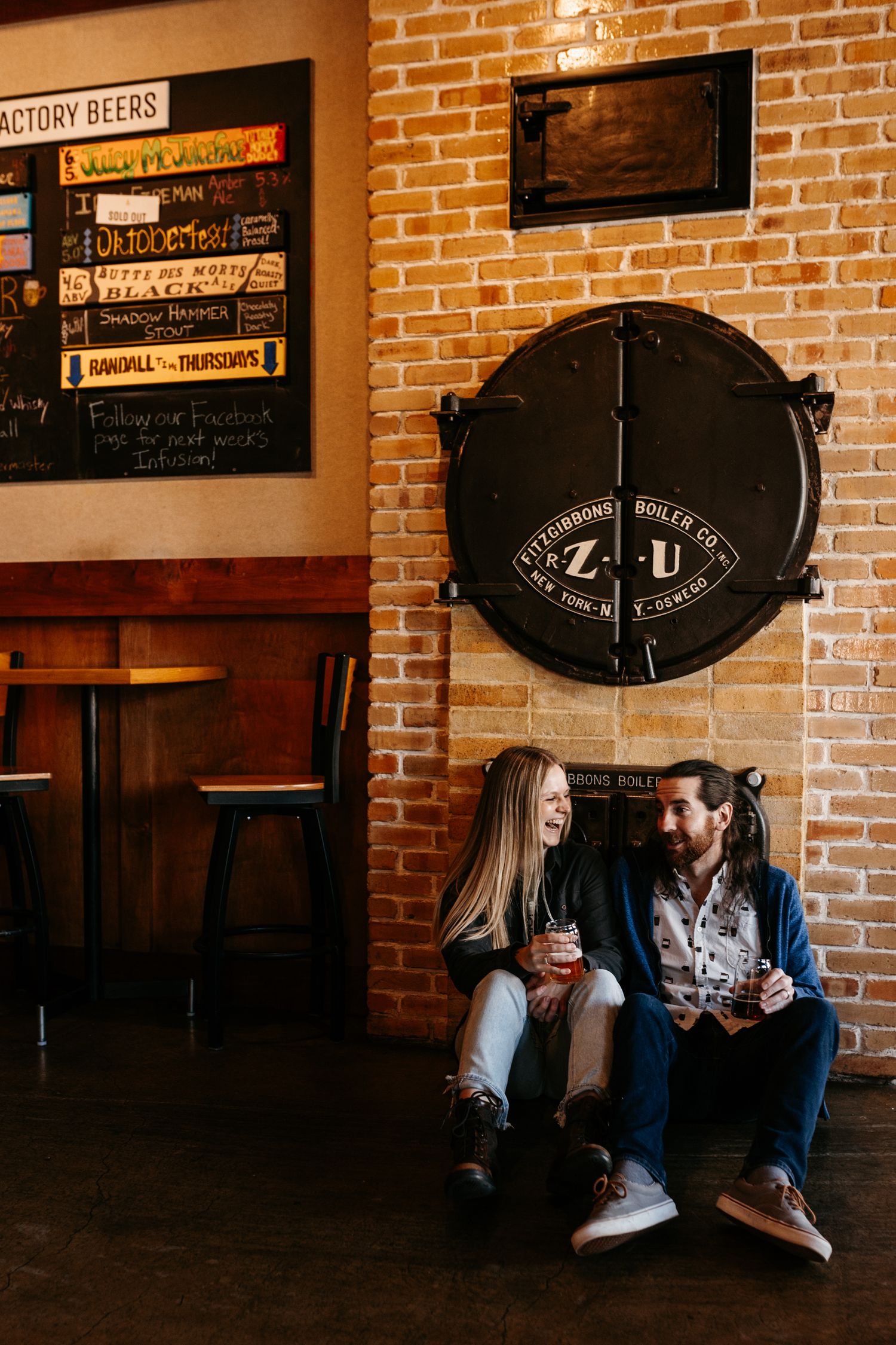 Two people enjoy craft beers while sitting against a rustic brick wall with brewery signage.