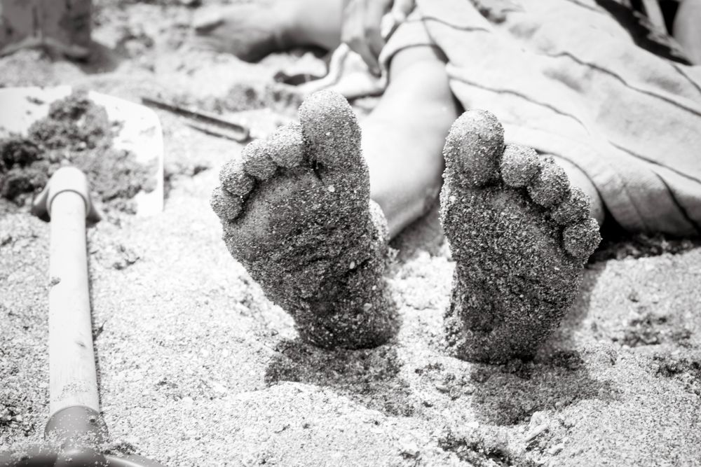Close-up of sandy feet sticking up from a child lying on a beach towel, captured in black and white with beach toys nearby.