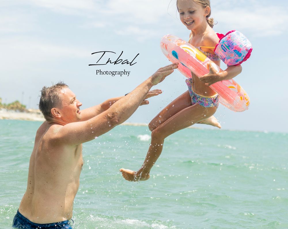 A father joyfully tosses his smiling daughter into the air at the beach, both enjoying a sunny day in the ocean waves.
