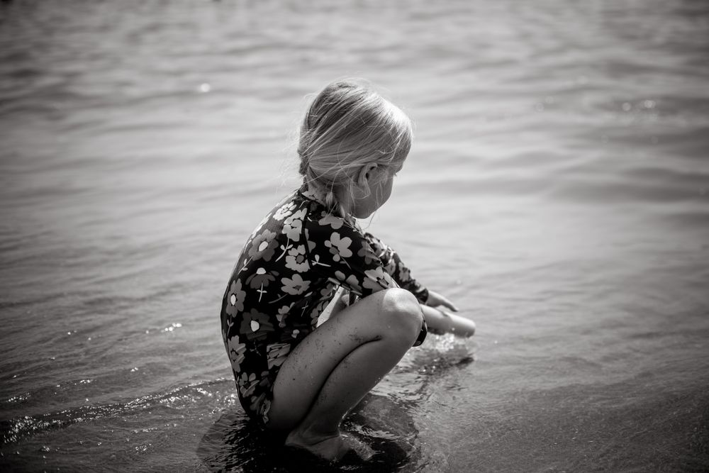 A young girl in a floral swimsuit crouches at the shoreline, quietly playing in the shallow water on a calm beach day.