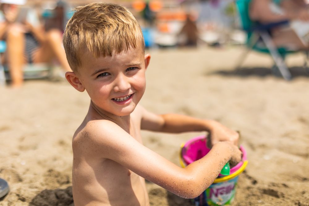 Smiling young boy plays with a colourful bucket in the sand on a sunny beach, surrounded by blurred beachgoers in the background.