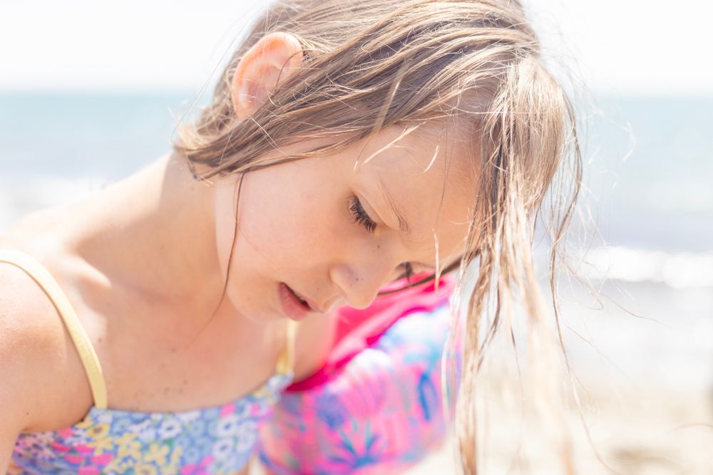 A young girl with wet hair gazes down thoughtfully at the beach, sunlight highlighting her face and pastel swimsuit.