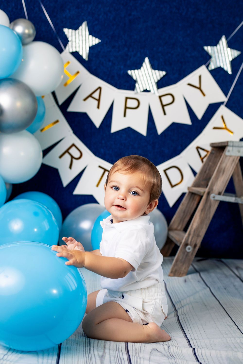 Birthday party scene with white and blue balloon arch, wooden ladder, and 'Happy Birthday' banner.