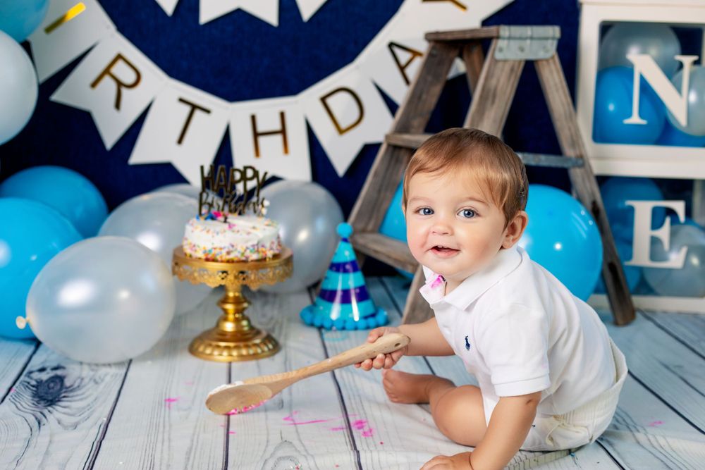 Birthday setup with gold cake stand, party hat, and blue balloon decorations on wooden floor.