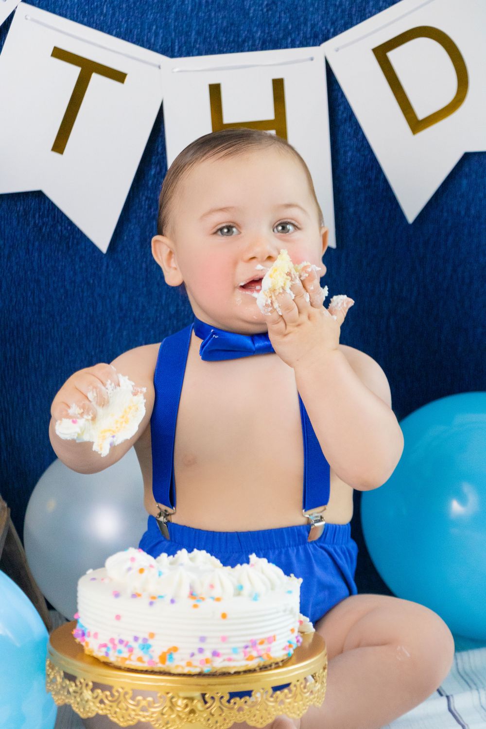 Birthday celebration with cake smash featuring blue suspenders outfit and white banner decorations.