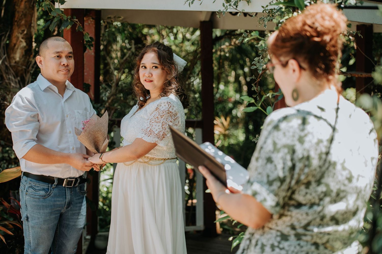 An intimate outdoor wedding ceremony takes place under trees with a couple exchanging vows.