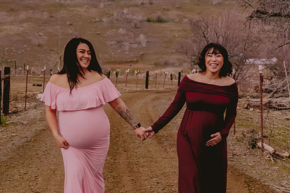 Two expectant mothers in pink and burgundy dresses hold hands on a dirt path against a desert backdrop.