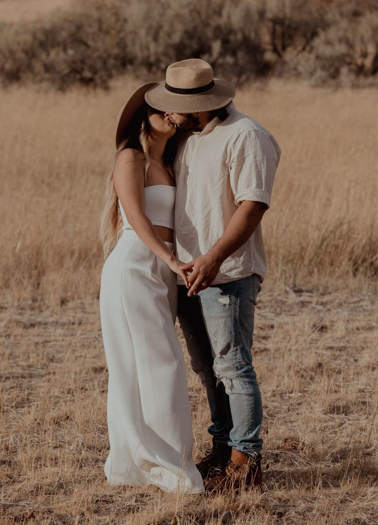 A romantic couple embraces in a golden field during sunset, with one wearing a brown hat and white outfit.