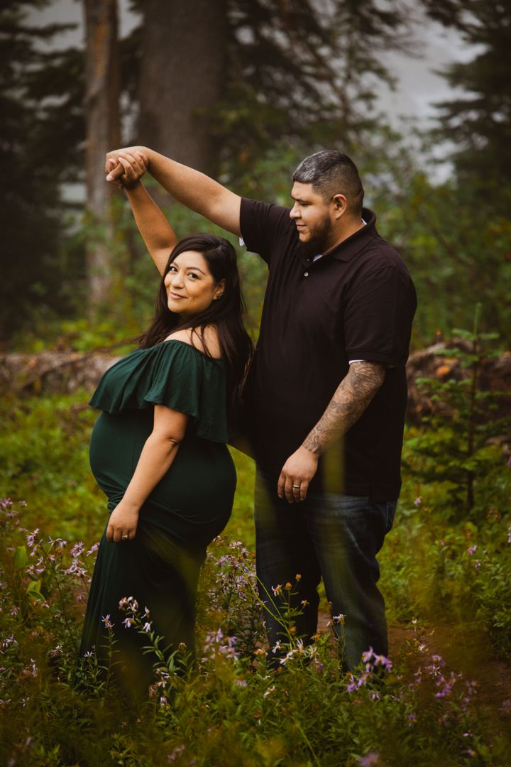 A couple poses playfully in a forest meadow filled with wildflowers during a maternity photoshoot.