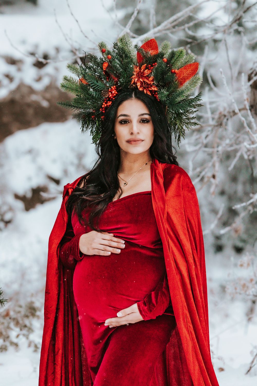 A maternity photoshoot featuring a red velvet dress and evergreen crown in a snowy setting.