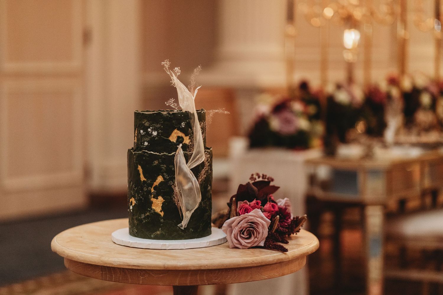A dark green wedding cake decorated with gold details and pink flowers on a marble table.