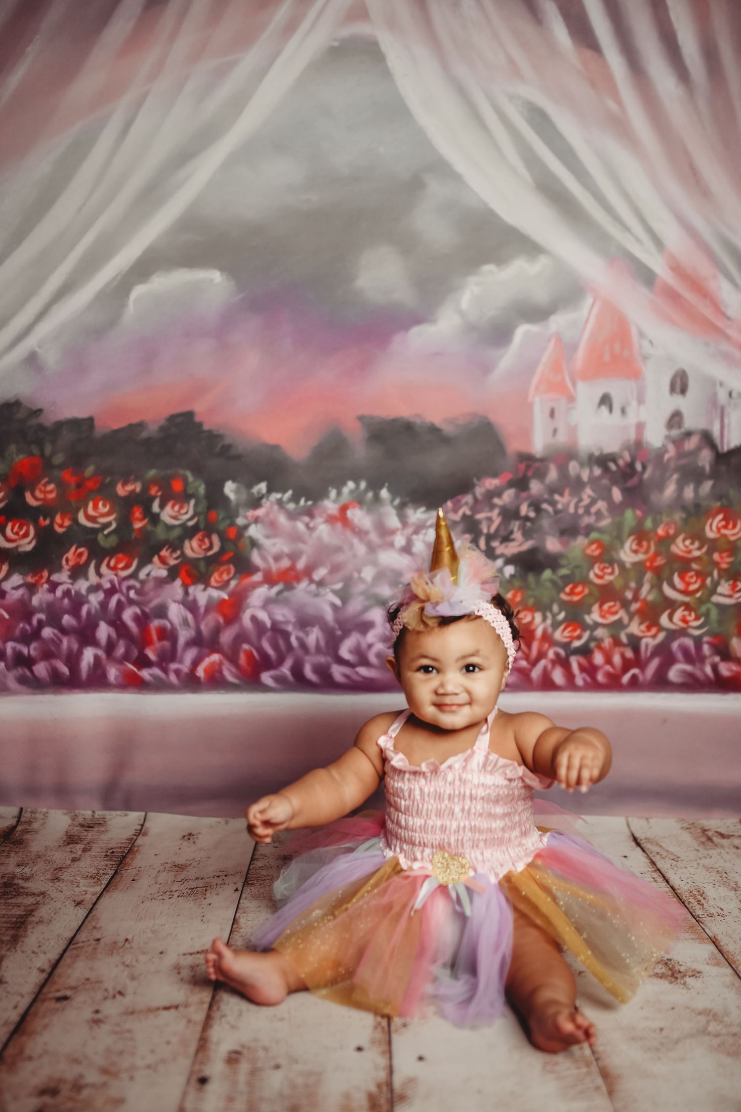 Baby wearing pink tulle dress lying on wooden floor with colorful floral backdrop.