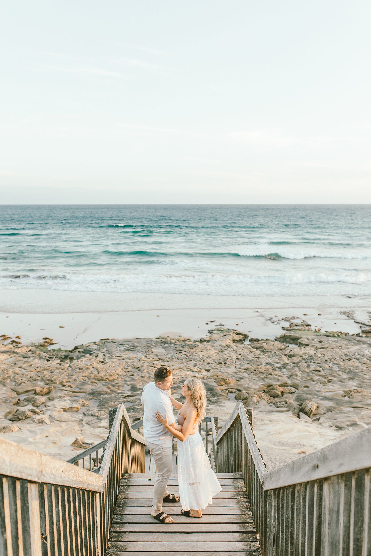 A couple stands on wooden stairs leading to a rocky beach with turquoise ocean waves.