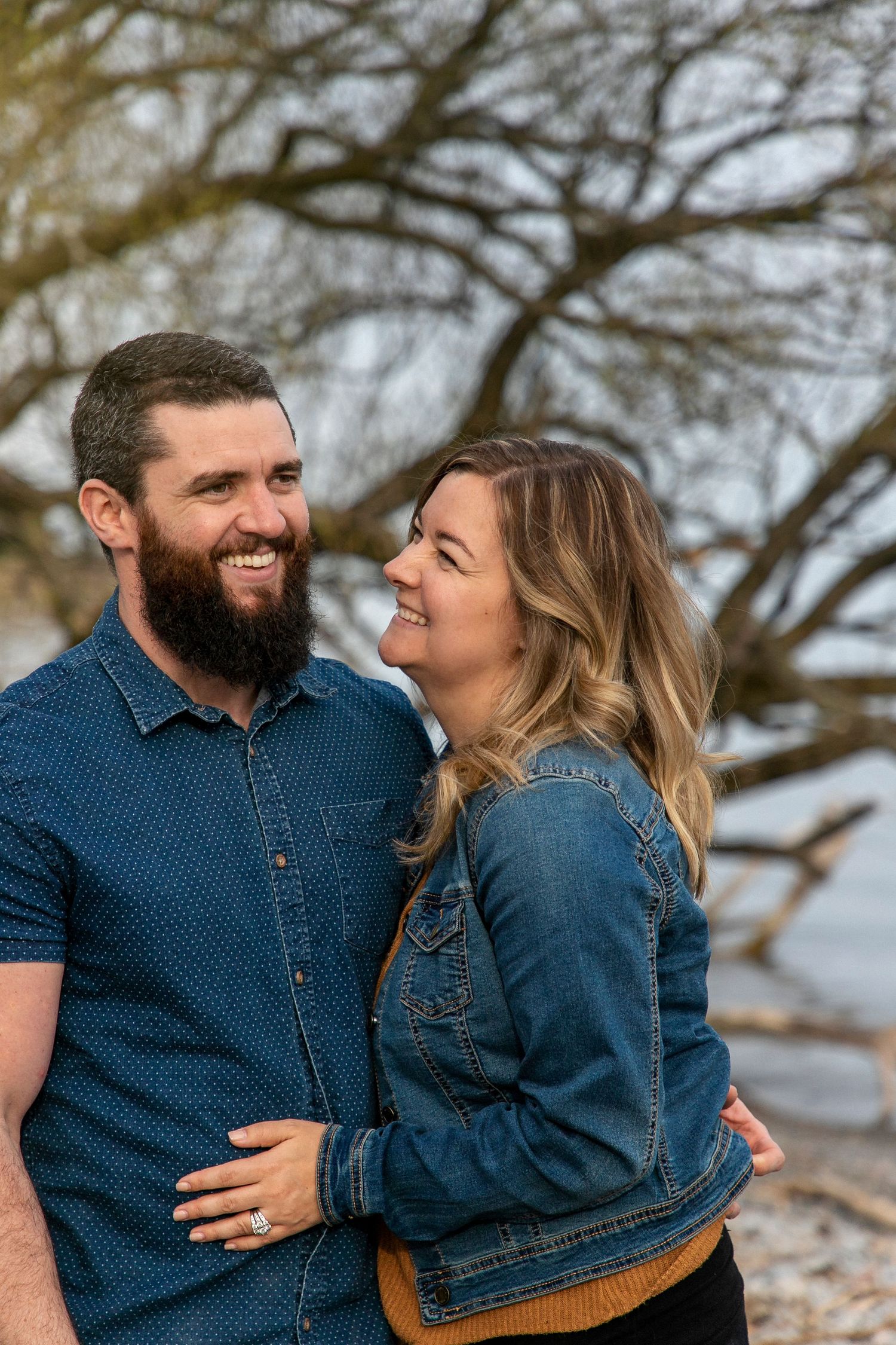 a couple cuddled up smiling and laughing at the beach with a tree behind them