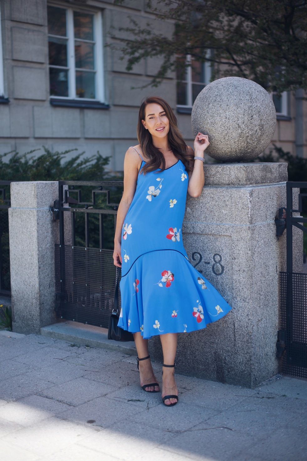 Woman in blue floral dress posing next to stone architectural detail on city street.
