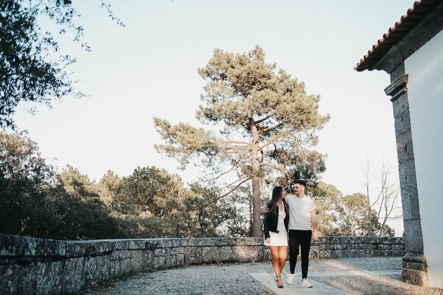 Couple walking along stone wall path with pine trees in soft daylight.