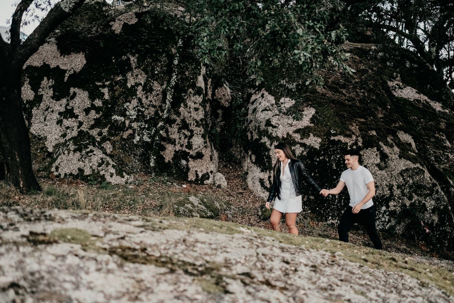 Couple walking along rocky terrain with dramatic dark trees in background.