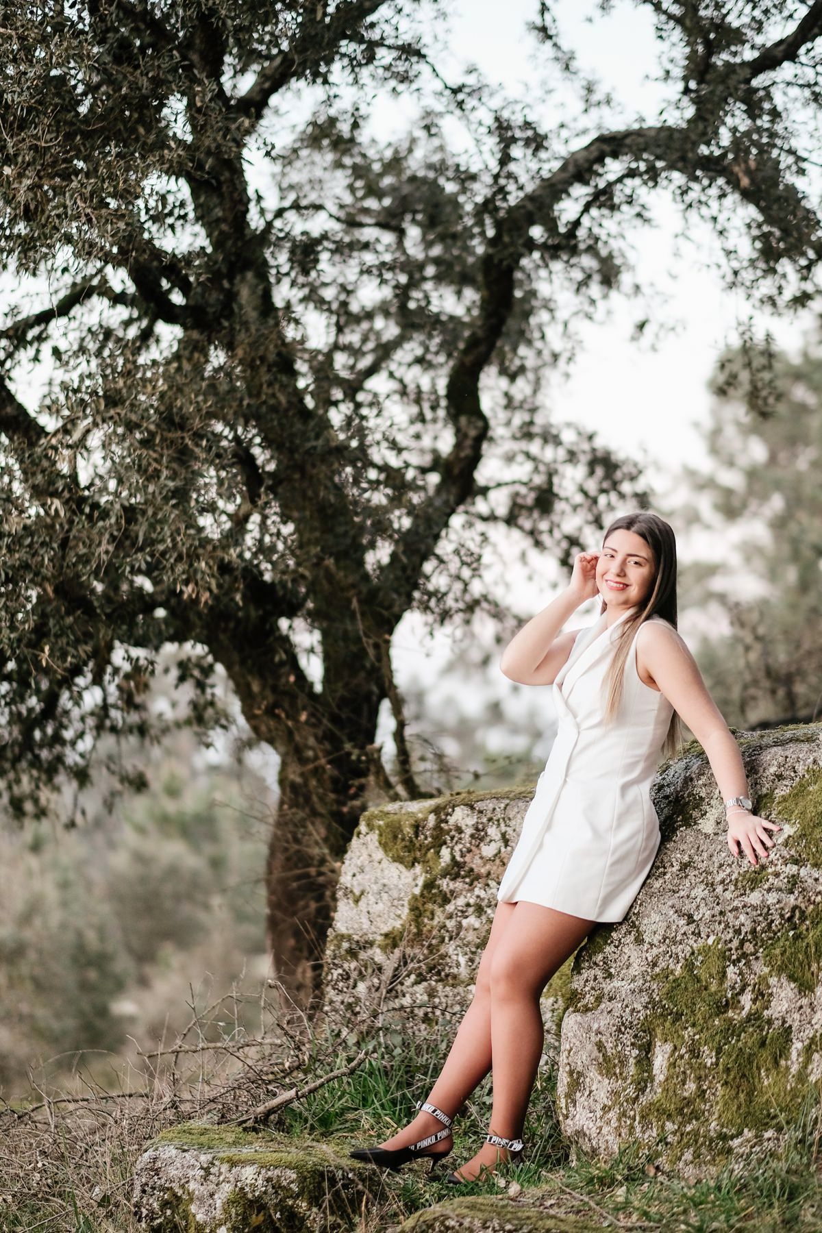 Woman in white dress posing against large tree on rocky terrain.