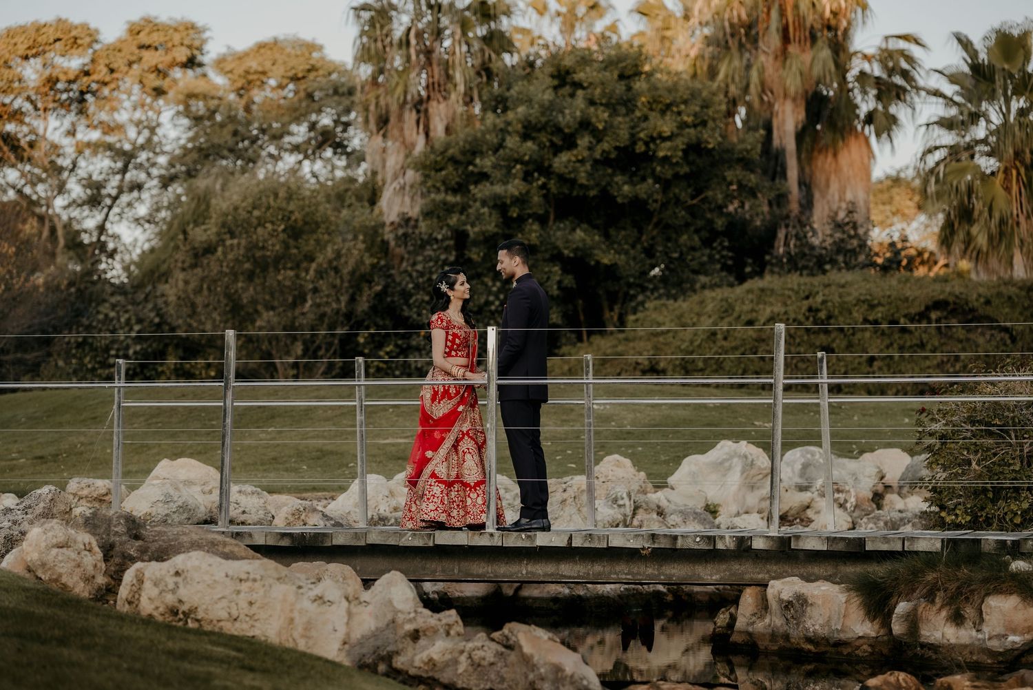 Couple in formal attire stands together on rustic bridge over rocky landscape.