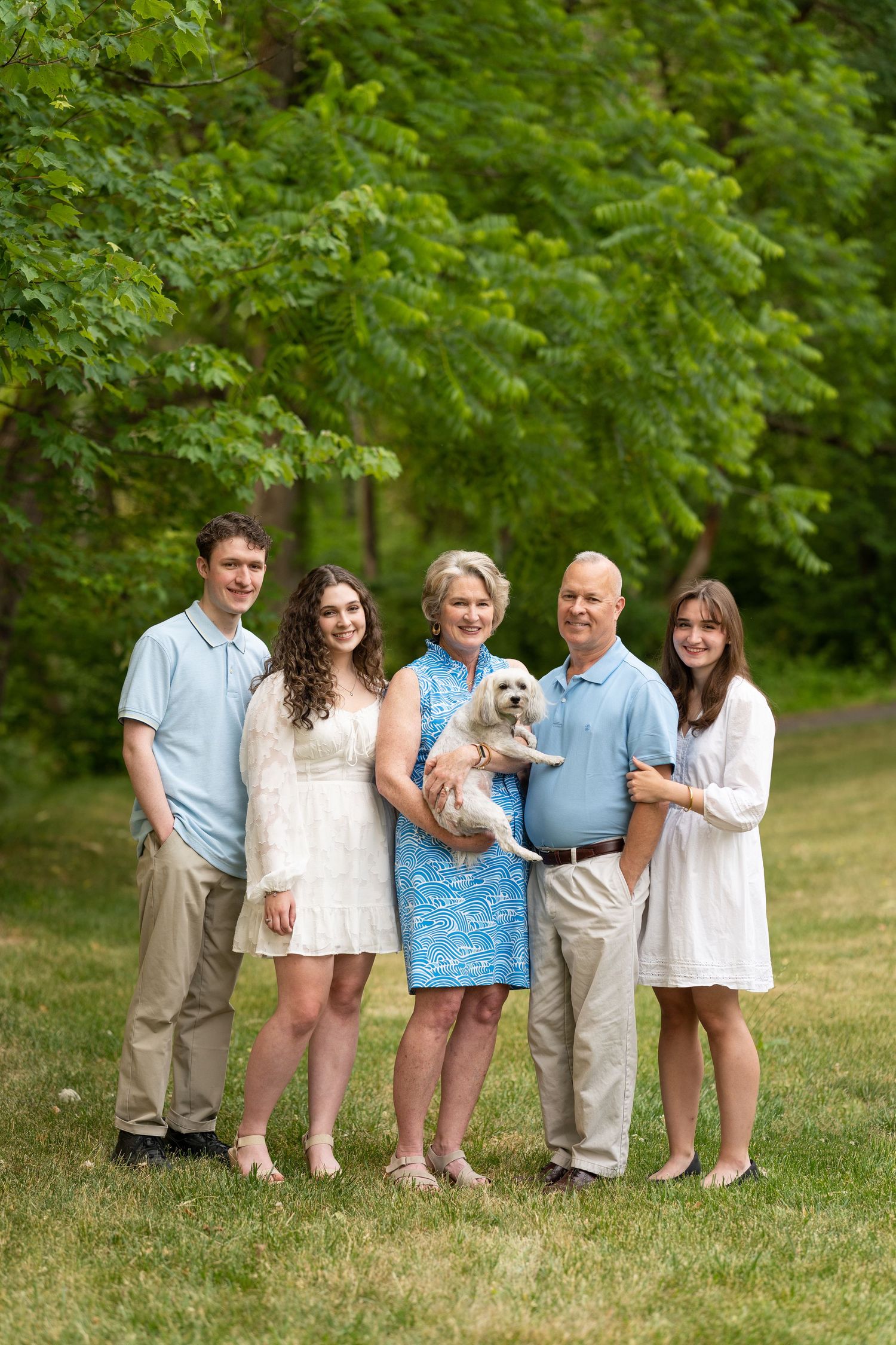 Family group with small white dog posing together on grassy lawn with green trees.