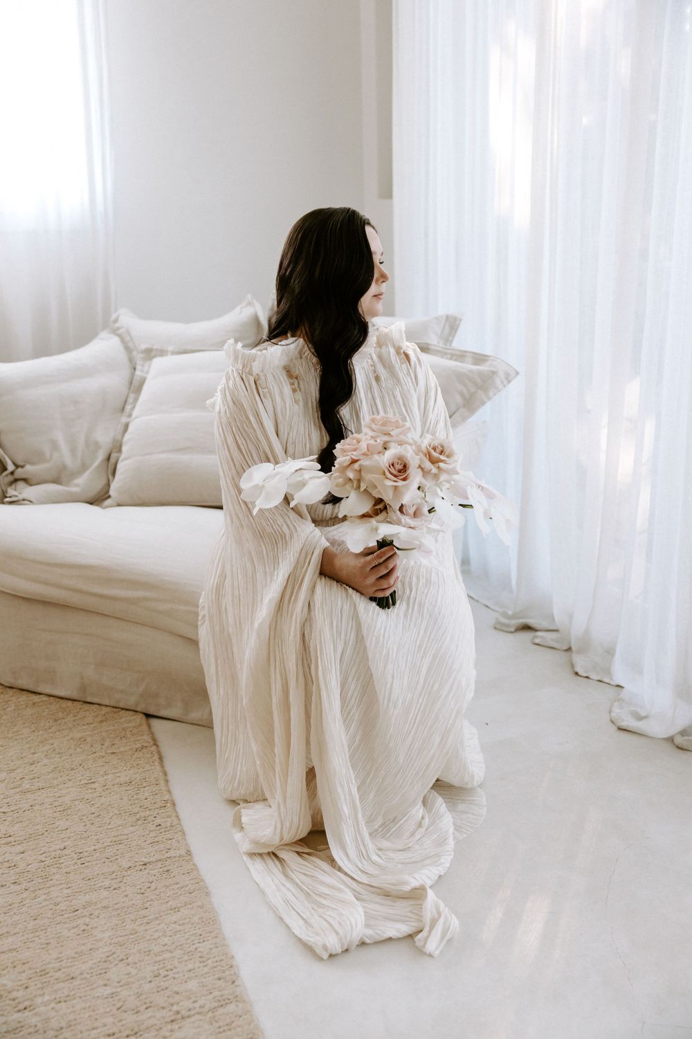 Ethereal scene with flowing white dress and delicate bouquet near bright windows.