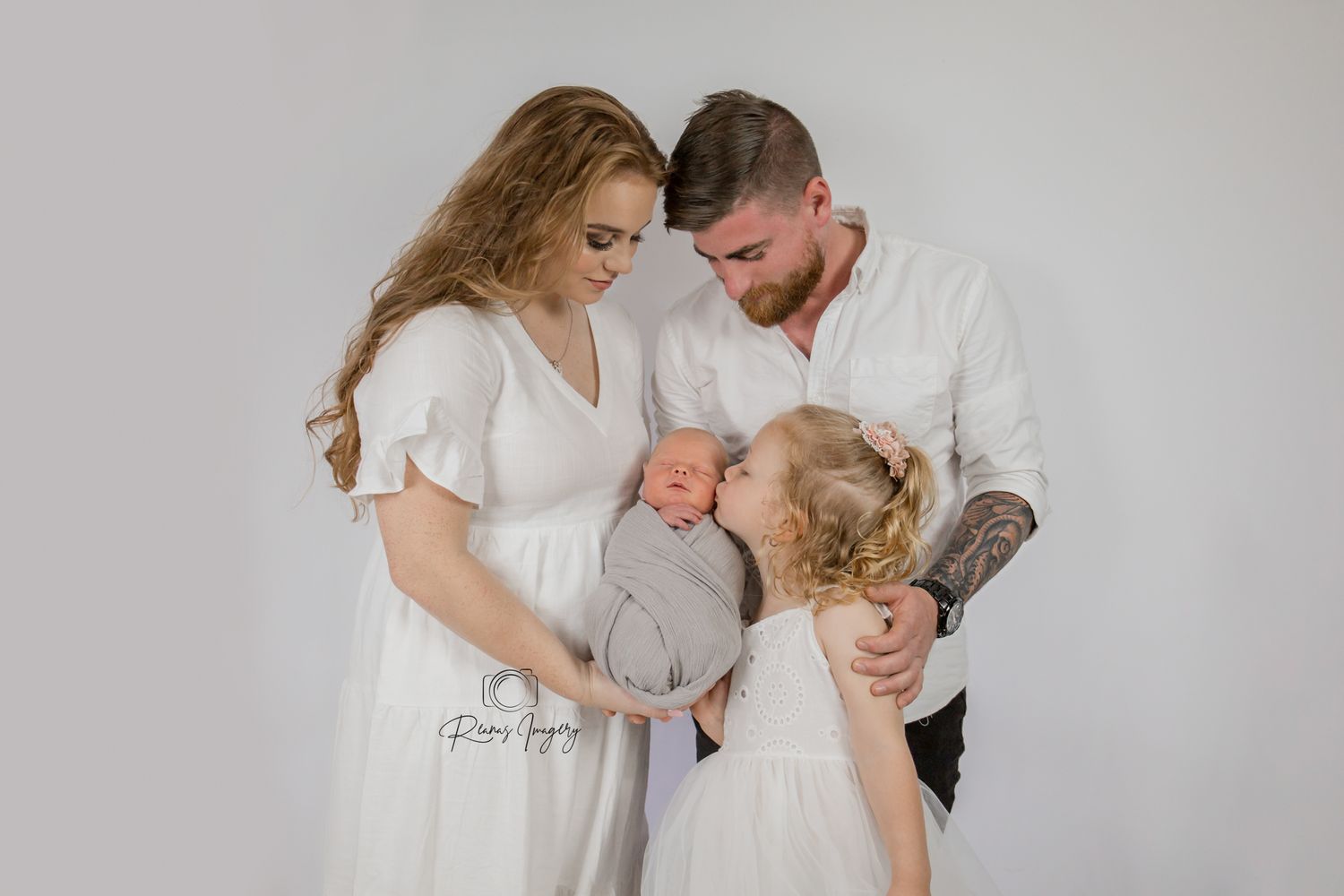 Parents in white outfits tenderly gaze at their swaddled newborn while young daughter looks on.