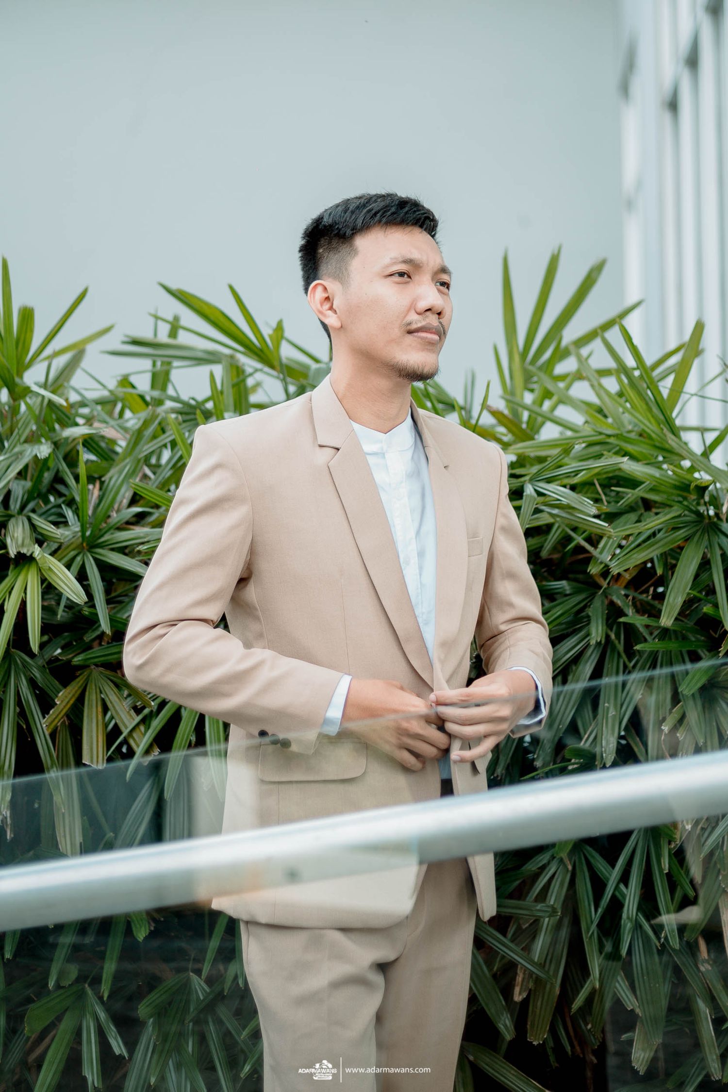 Man in beige suit standing by modern glass railing with tropical plants.