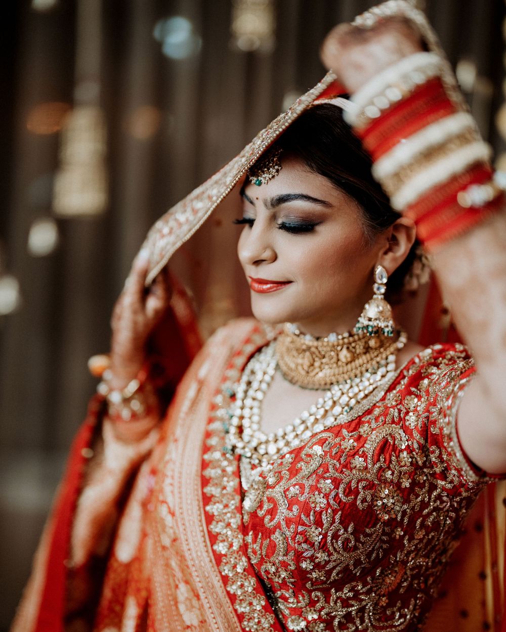 Traditional Indian bride in red lehenga with intricate embroidery and jewelry poses elegantly in soft lighting.