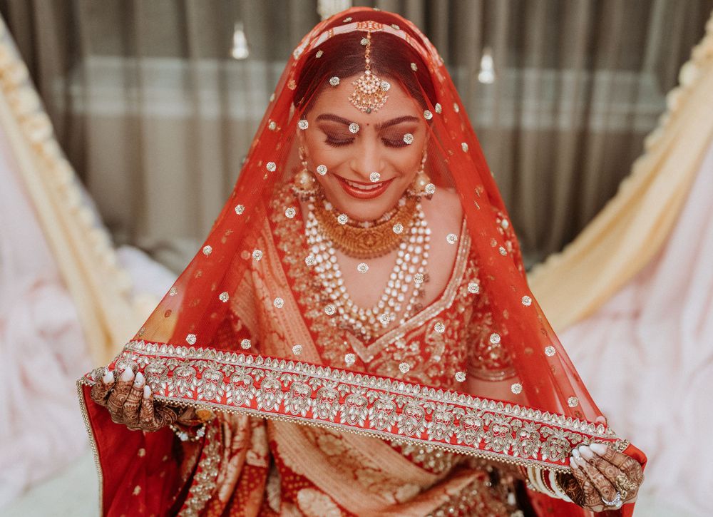 Traditional Indian bride in red lehenga with ornate jewelry and veil smiles while holding decorative accessories.