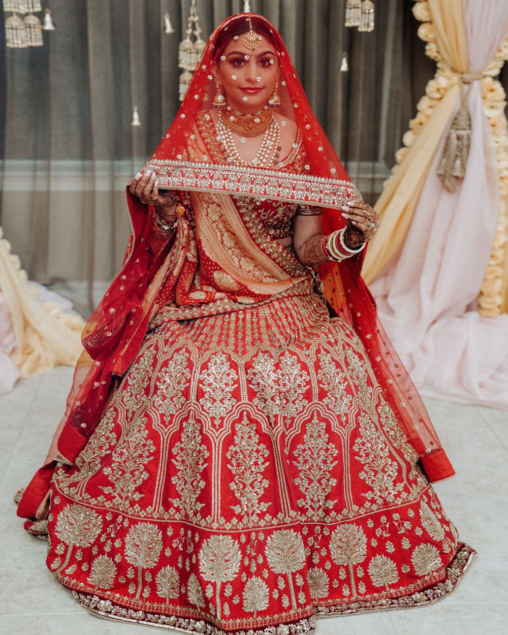 Bride wearing ornate red and gold bridal lehenga with detailed embroidery holds traditional dupatta.