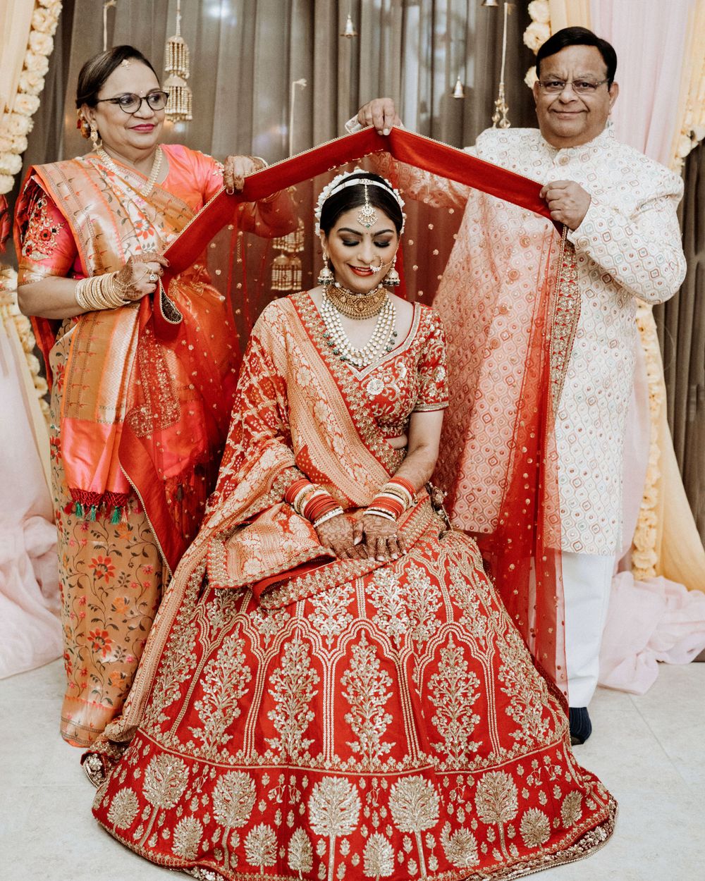 Traditional wedding ceremony scene with family members holding red dupatta over seated bride.