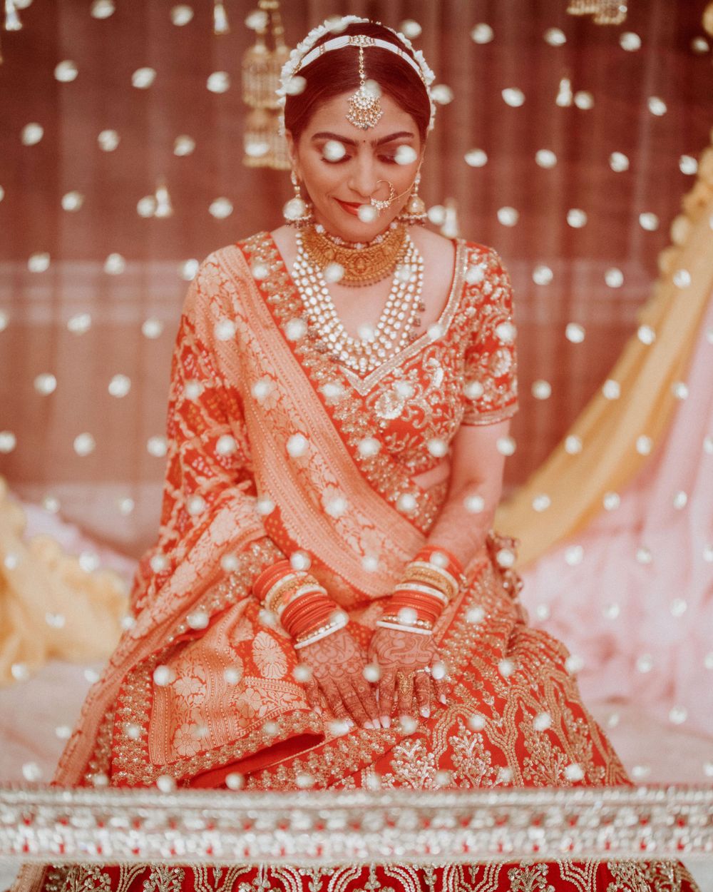 Bride in red and gold lehenga sits with decorated henna hands against sparkly backdrop.